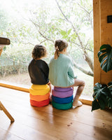 Two children sitting on colorful Stapelstein original rainbow inside a room with a large window view of trees.