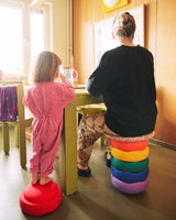 A child in a pink jumpsuit stands on a red Stapelstein while an adult sits on a stack of colorful Stapelstein Rainbow stones at a wooden table in a cozy, sunlit room.