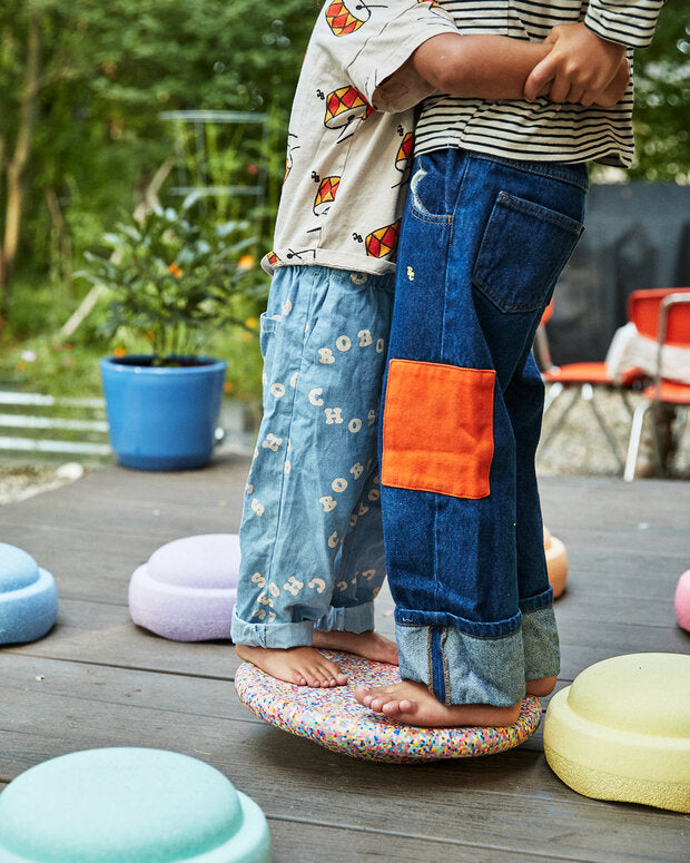 Two children standing on a colorful balance board outdoors with a garden background.