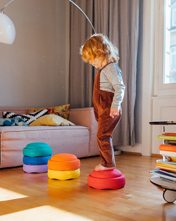 Child standing on a colorful Stapelstein rainbow classic in a room with a sofa and books.
