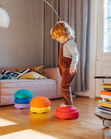 Child standing on a colorful Stapelstein rainbow classic in a room with a sofa and books.